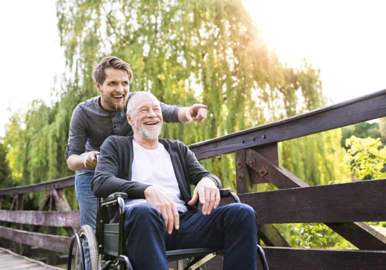 Hipster,son,walking,with,disabled,father,in,wheelchair,at,park.