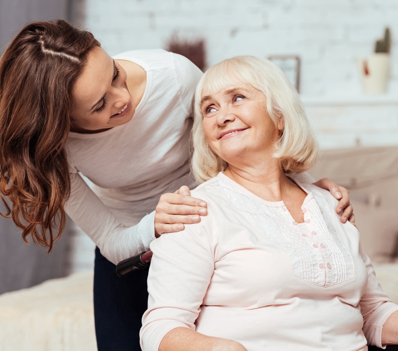 Young female caregiver placing hands on shoulders of happy senior woman in wheelchair
